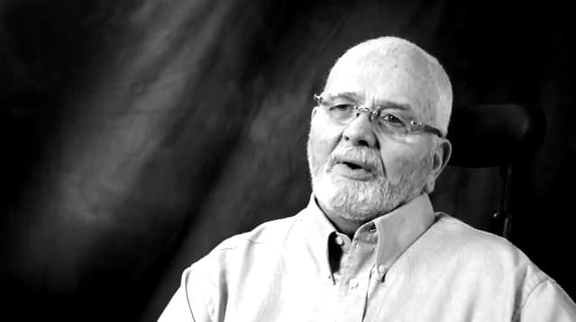 An older man with a beard and glasses, wearing a shirt emblazoned with "Sun Valley Construction," is seated and appears to be in mid-conversation against a dark, textured background.