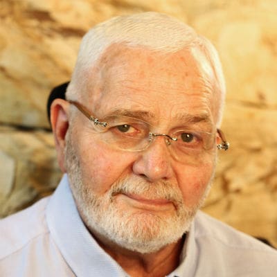 An elderly man with gray hair and a beard, wearing glasses and a light blue shirt emblazoned with the Sun Valley Construction logo, poses for a photograph in front of a stone wall background.