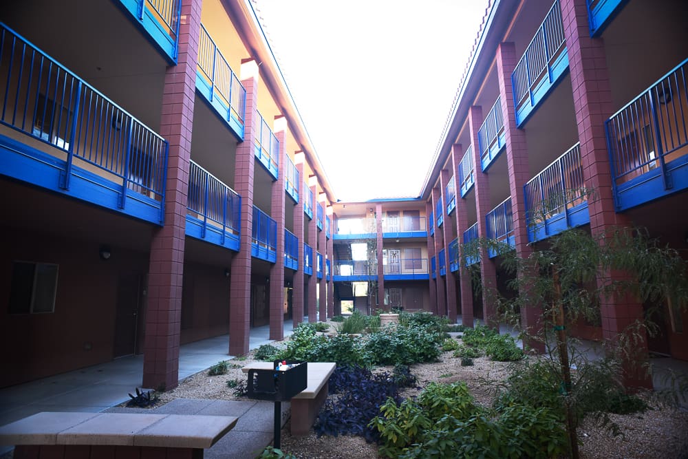 An empty courtyard within a residential building featuring two stories with blue railings and red exterior walls, surrounded by small green shrubs and trees under a bright sky.