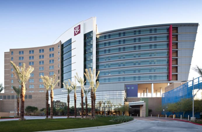 A modern hospital building with palm trees in the foreground and the medical center's logo displayed prominently on the side of the structure.