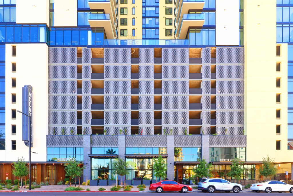 Modern multi-story building facade with patterned balconies, ground-level retail stores, and parked cars in front.