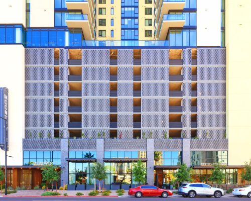 Modern multi-story building facade with patterned balconies, ground-level retail stores, and parked cars in front.