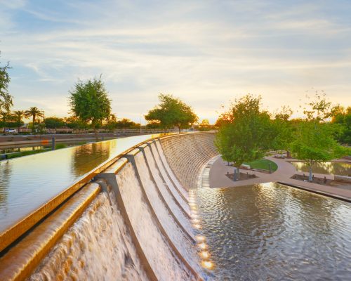 Sunset over a tranquil urban canal with flowing water, lighted paths, and lush greenery on both sides.