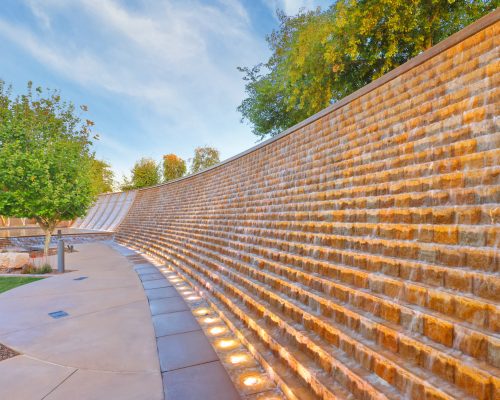 A large illuminated stone retaining wall along a curved pathway in a landscaped park, with trees and a clear sky visible.