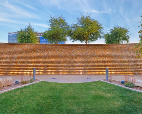 Illuminated water wall in a tranquil outdoor park setting with trees and a lush green lawn at dusk.