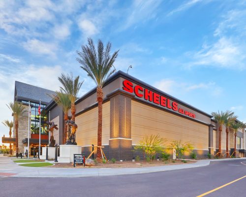 Exterior view of a scheels store featuring prominent signage, a statue of a sports figure, and a clear blue sky at sunset.