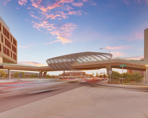 Modern cityscape at twilight showing a distinctive circular pedestrian bridge over a busy street, with light streaks from moving vehicles and colorful clouds above.