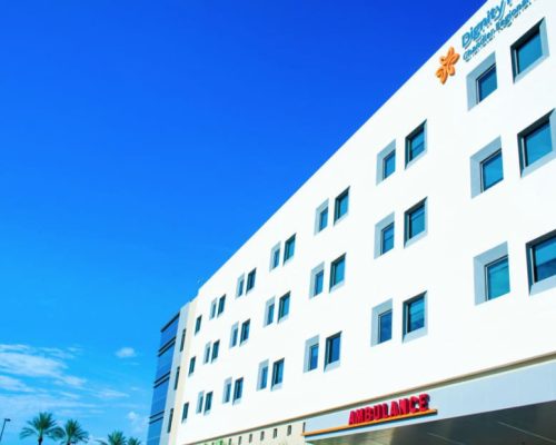 A modern hospital building under a clear blue sky with the ambulance entrance visible on the ground floor.