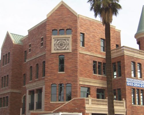 A red brick building with a conical tower, decorative stonework, and multiple arched windows, located under a partly cloudy sky.