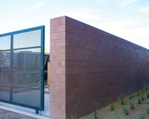 A sturdy metal gate set within a high, textured brown wall, leading to a utility area with various equipment, under a clear blue sky.