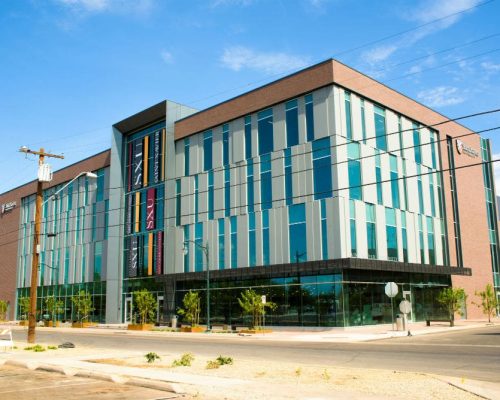 Modern multi-story building with glass facade and brick elements on a sunny day, with power lines and a clear blue sky in the background.