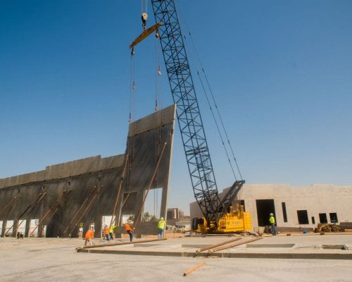 A large crane hoisting a concrete panel at a construction site with workers overseeing the process.