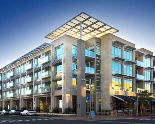 Modern multi-story residential building with large windows and balconies, captured during dusk with street parking in the foreground.