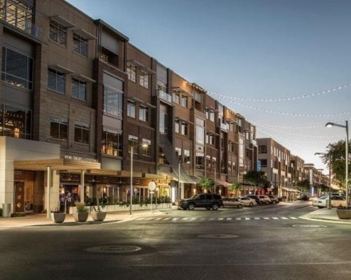 A tree-lined urban street at dusk with strings of lights above, flanked by modern residential buildings and some parked cars.